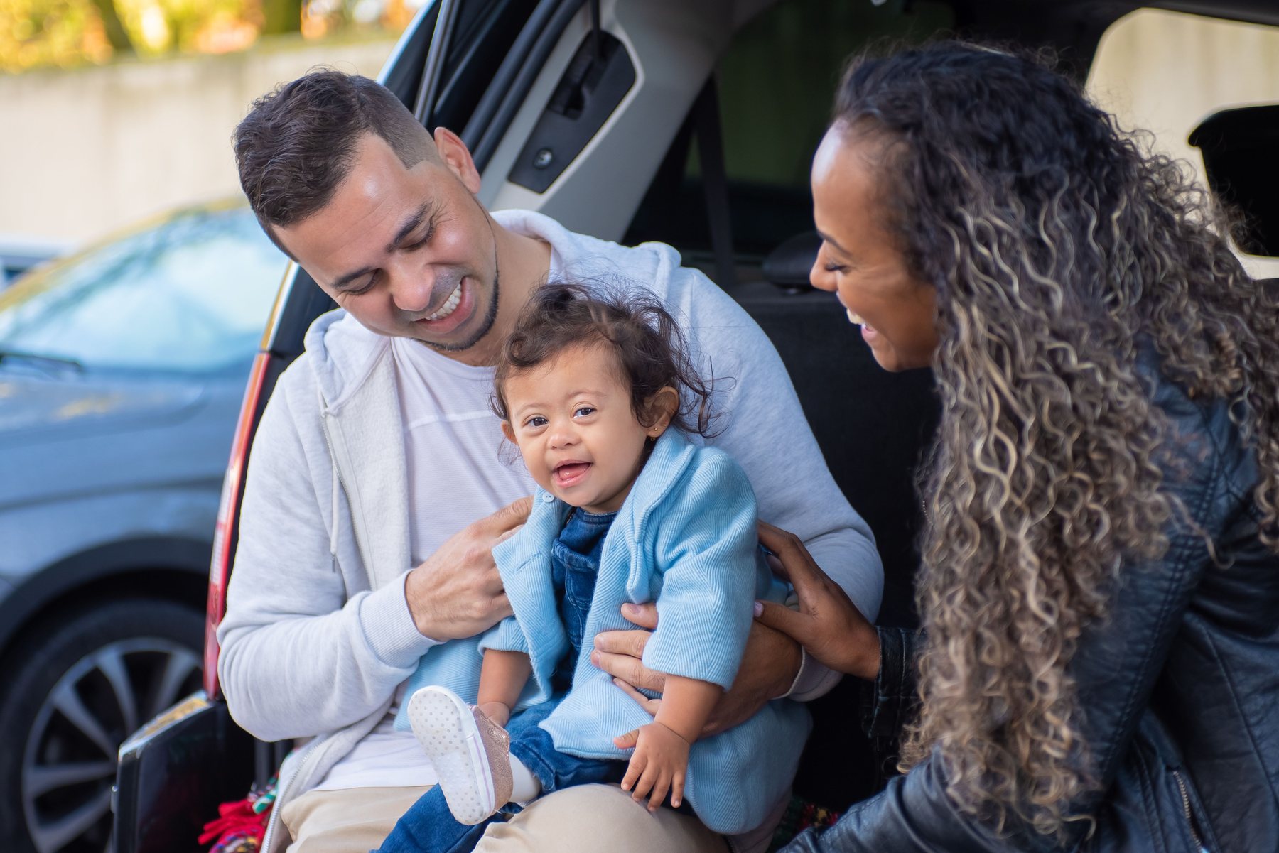 Parents laughing with their toddler, sitting in the open trunk of their car