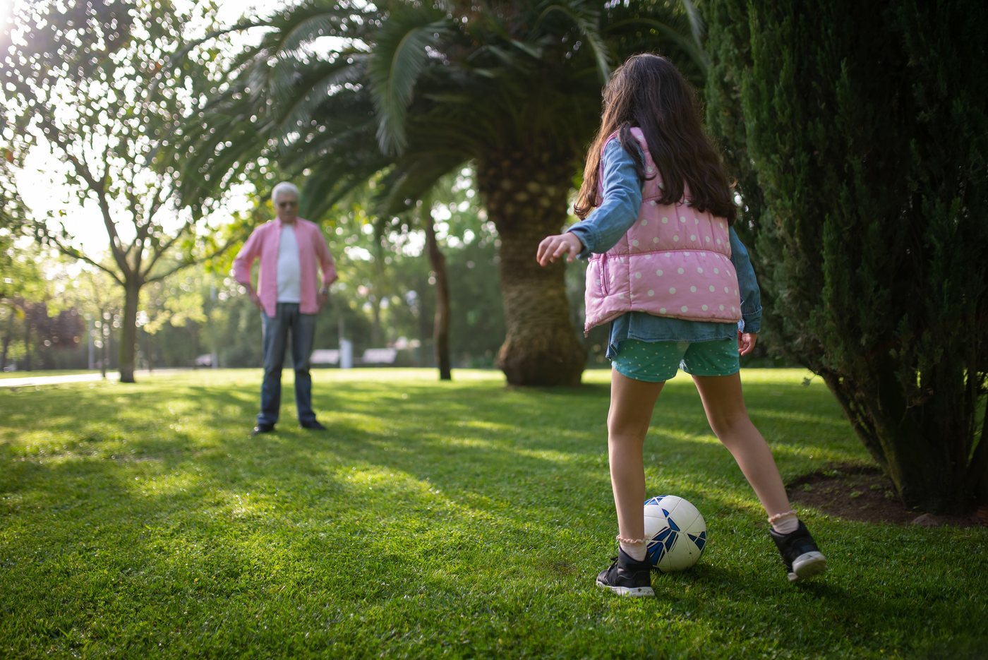 A girl kicking a ball to her grandfather on a green lawn under palms