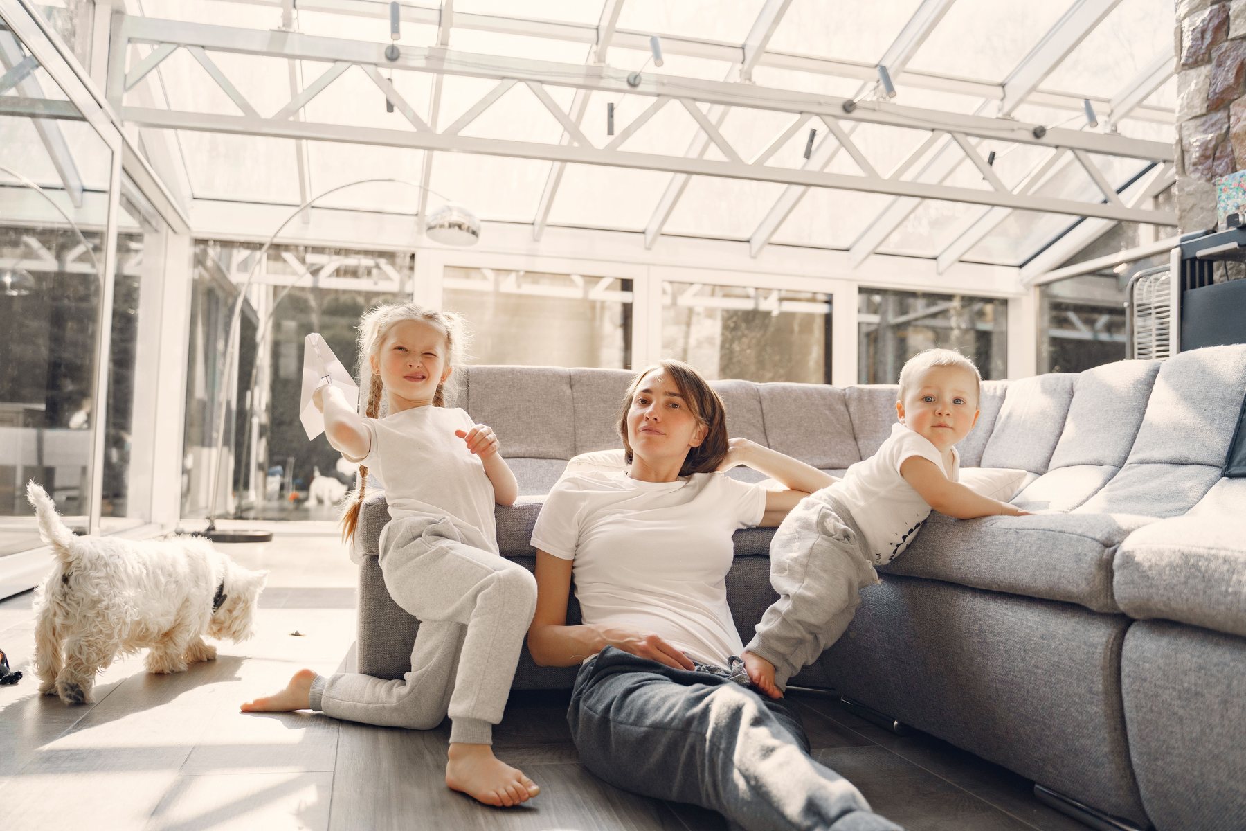 A mother, two children and a small white dog on the floor of a bright sunroom